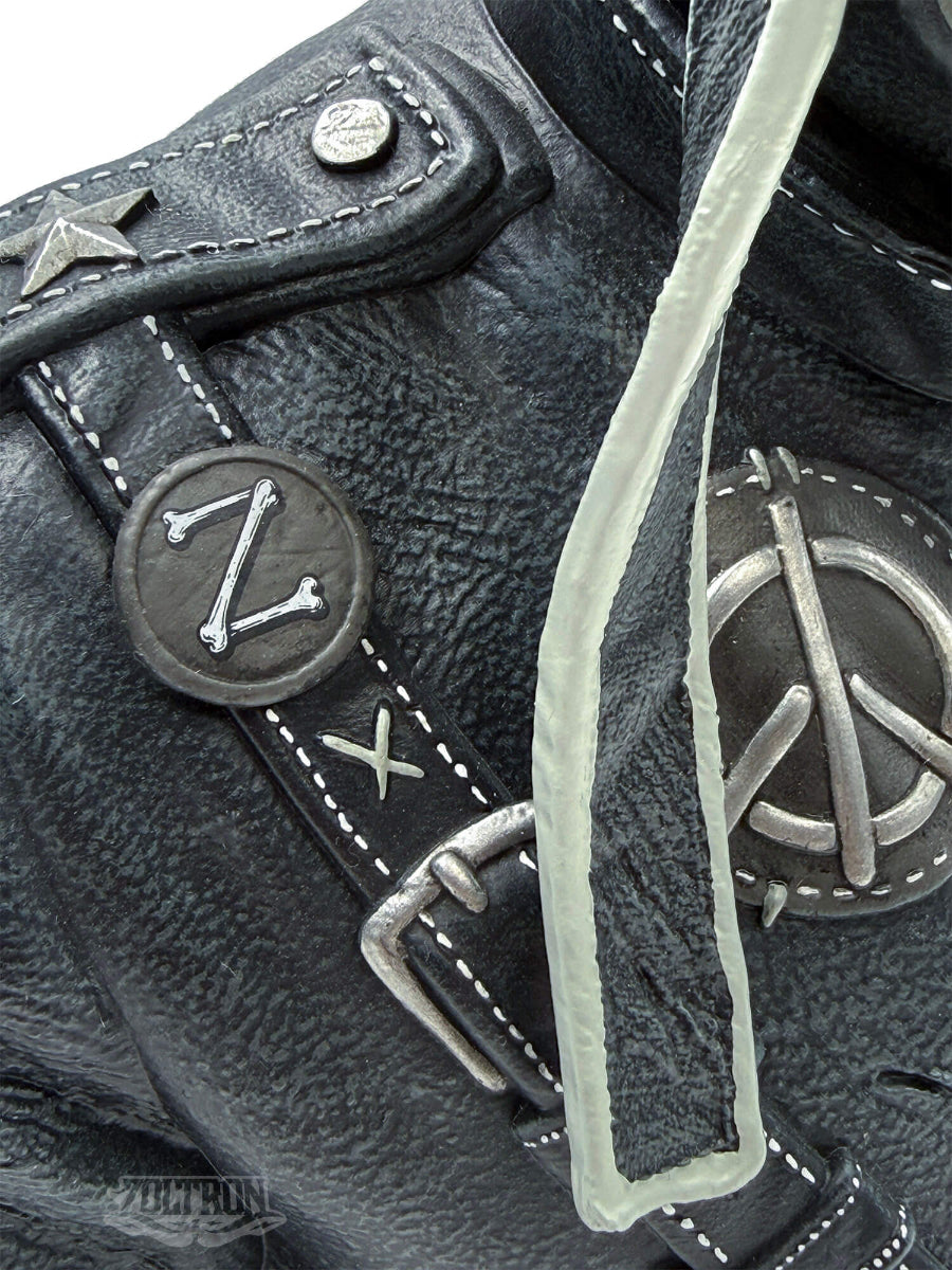 Close-up of a detailed Zoltron artist figure sculpture, black leather jacket strap with white contrast stitching, a silver metal buckle, and a circular black button featuring a white bone-style letter Z. A silver peace sign patch is visible on the adjacent section.