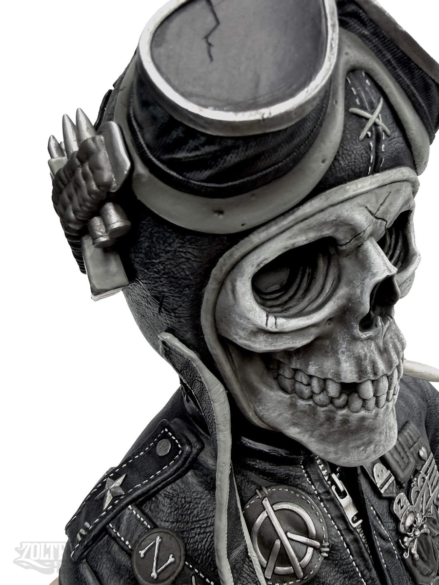 A close-up of a highly detailed resin sculpture by artist Zoltron, featuring a highly detailed skull figurine wearing a cracked leather aviator helmet, a weathered black leather jacket, war medallions and adornments, smoking a cigarette, set against a plain white background.